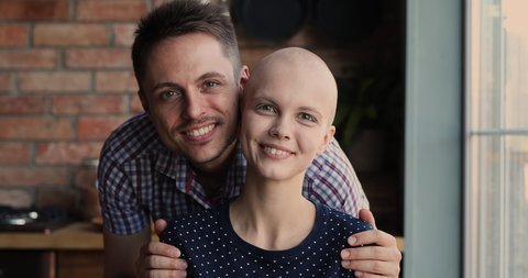 Affectionate young man cuddling shoulders of smiling hairless 25s woman, supporting beloved wife during oncology cancer disease treatment, feeling hopeful looking at camera, struggle for life concept.