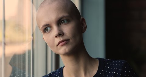 Head shot dreamy young woman with bald head standing near window, breathing fresh air, finding strength for fight with cancer, oncology treatment center hopeful female patient meditating indoors.