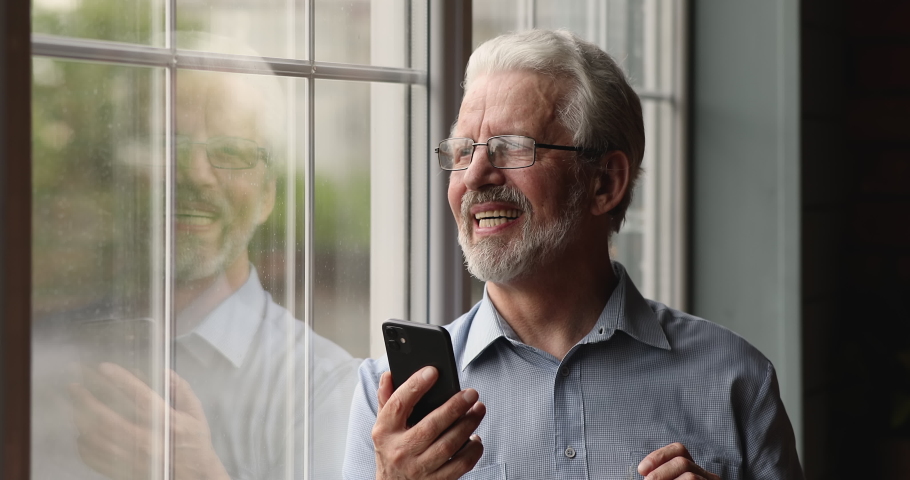 Dreamy old senior man in eyeglasses standing near window, enjoying using software applications on cellphone, reading message or email with good news, elderly generation people and modern technology. | Shutterstock HD Video #1071403411