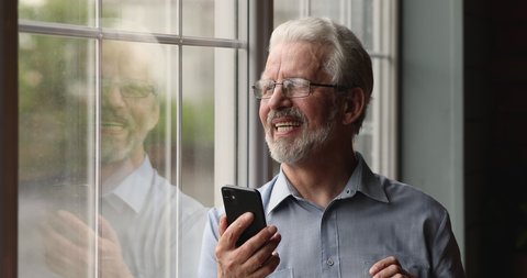 Dreamy old senior man in eyeglasses standing near window, enjoying using software applications on cellphone, reading message or email with good news, elderly generation people and modern technology.