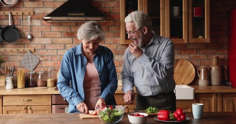 Happy loving middle aged senior family couple talking chatting, enjoying cooking together in old-fashioned kitchen, chopping fresh vegetables, preparing healthy food dinner together on weekend at home
