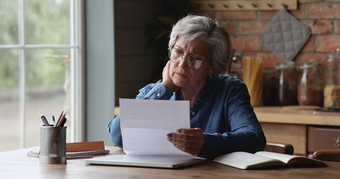 Curious older mature retired woman in glasses opening envelope with correspondence, reading paper letter, feeling confused of getting unpleasant news, considering banking notification indoors.
