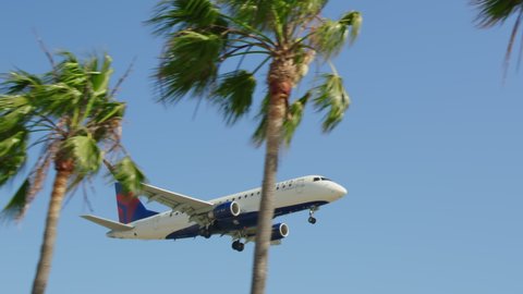 Los Angeles airport, USA, April 2021. Summer California travel. Aircraft arriving at airport, RED shot. Slow motion landing airplane behind cinematic green palm tress with clear blue sky on background