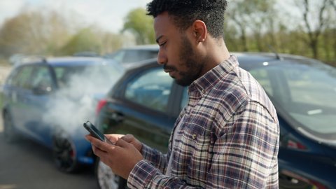 Young man standing by damaged car after traffic accident reporting incident to insurance company using text message on mobile phone - shot in slow motion