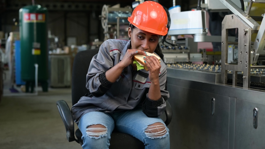A young black woman in an orange helmet eats a sandwich in a production facility, next to a conveyor belt. Production worker taking a lunch break at the workplace