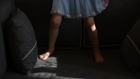 Toddler girl in short blue tutu skirt with barefoot legs jump on couch in slow motion. Kid having fun at home. Authentic lifestyle concept and happy childhood
