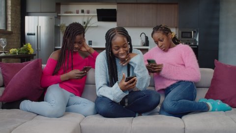 Addicted to cellphone lovely african american teenage girls sitting on couch, phubbing each other, browsing social media content online on smart phones, showing ignorance and apathy indoors.