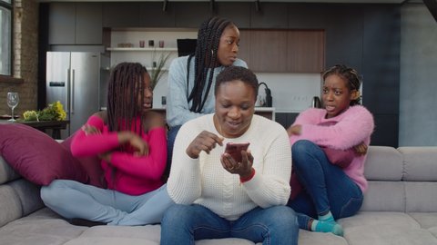 Addicted to smartphone carefree beautiful african mother browsing social media content online on cellphone, ignoring cute upset disappointed teenage daughters while family relaxing in home interior.