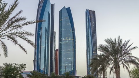 Skyscrapers of Abu Dhabi illuminated with Etihad Towers buildings day to night transition timelapse. Palms in park near by. United Arab Emirates
