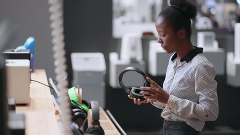 young afro-american woman is putting headphones in trading hall of appliance shop, testing new model before buying