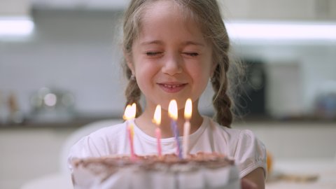 Close-up of charming little girl with closed eyes making wish and blowing candles on birthday cake. Portrait of happy Caucasian pretty child celebrating birthday indoors at home. Joy and lifestyle