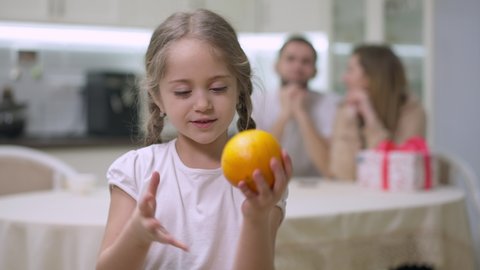 Cheerful girl juggling orange and smelling delicious fruit looking at camera smiling. portrait of happy joyful Caucasian child posing with healthful dessert and blurred couple of parents at background