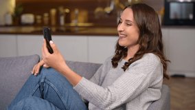 Video call concept. Young freelance woman waving hello into phone webcam, cheerful female connected via video with family, friends or colleagues lying down on the couch at home