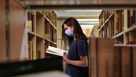 Student Wearing Face Mask Reading Book Inside Library Zoom In. Young man student with long hair is wearing face mask protection and reading a book inside a library. Zoom in