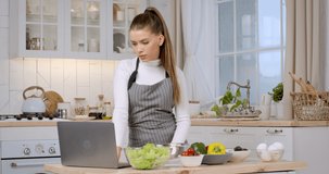 Online culinary courses. Young caucasian woman looking at laptop and checking all products need for recipe, preparing lunch at kitchen, slow motion