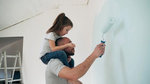 Caucasian man and girl paint the wall Daughter sits on the shoulders of her father and paints the wall with a roller Happy family makes repairs in the room Side view