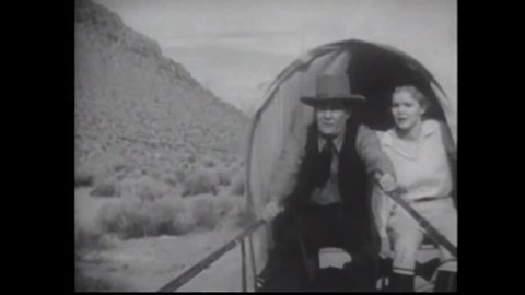 CIRCA 1934 - In this western film, a covered wagon with a pioneer man and woman inside is chased off a cliff into a lake by a posse.