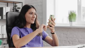 Close up of joyful smiling young woman tapping on mobile phone. Portrait of beautiful brunette girl texting on smartphone, indoor. Female laughing and uses cellphone in office. Girl look at the phone