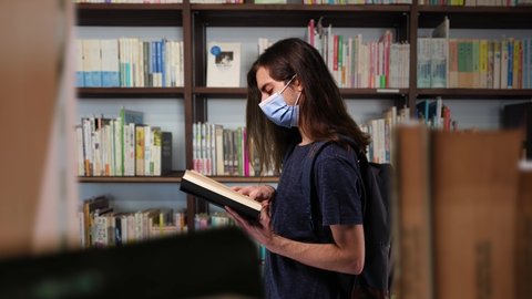 Student Boy Wearing Face Mask Reading Book Inside Library Zoom In. Young man student with long hair is wearing face mask protection and reading a book inside a library. Zoom in