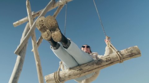 A cheerful young woman in a light jacket, blue jeans and sunglasses is swinging on a wooden swing. Cool colors. Bottom view slow motion. looks into the camera. Good mood 