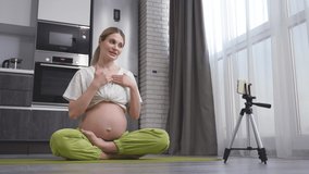 pregnant woman sitting in a lotus position on the floor, ready for prenatal yoga exercises, in sports clothes, at home alone, records a video lesson online