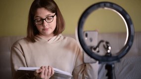 Girl book blogger with eye glasses recording vlog on digital smartphone cam at home. Young woman using ring light for register her book blog, book review