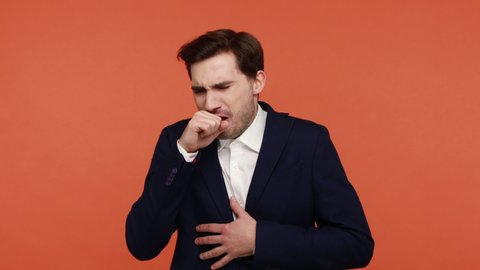 Seasonal flu. Unhealthy bearded man in official suit coughing covering mouth with hand, choke allergy or influenza symptoms, feeling unwell. Indoor studio shot isolated on orange background