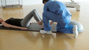 Female patient lying down on a mat, exercising with her physiotherapist using resistance band for legs, in a gymnasium or clinic. Physical therapy concept.