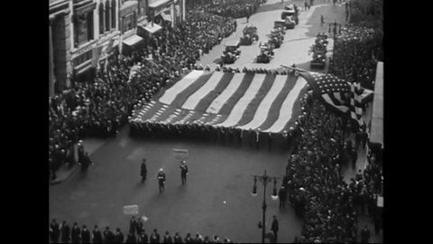 CIRCA 1919 - A giant American flag is carried in a patriotic parade in Manhattan, New York. Ships pass under the Brookyln Bridge on the East River.