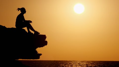 Silhouette of hipster woman puts headphones and enjoys music while sitting on cliff face looking at sea at sunset.