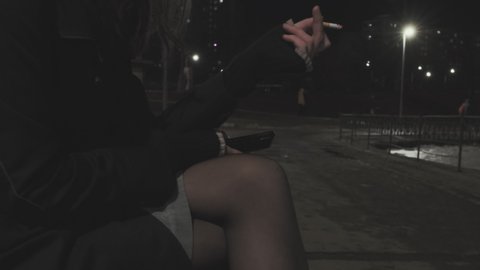 Girl in a mini skirt with long, black hair sits on a bench and smokes a cigarette. It night outside, lights are shining, people are walking in the background. cold weather