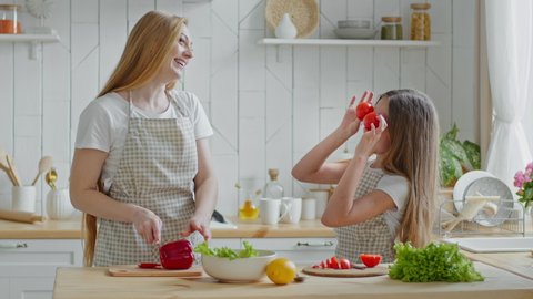 Adult mother housewife cuts red pepper vegetable ingredient salad daughter child teen girl helps mom with cooking holds two tomatoes instead of eyes posing laugh mommy woman having fun in home kitchen