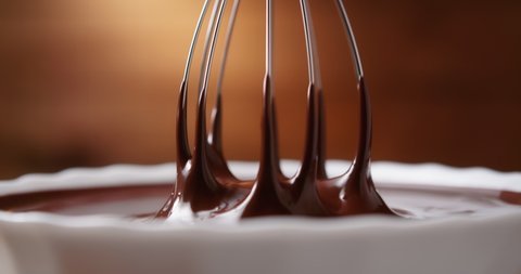 Close-up mixing melted liquid chocolate with steel whisk in kitchen. Hot chocolate mix swirl and drip in white bowl on wooden background. Cooking handmade chocolate dessert. Confectionery concept