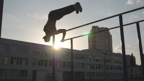 Silhouette of male gymnast doing push ups in handstand on uneven bars outdoor. Young athlete doing stunts on parallel bars. Strong muscular man showing performance at sport ground. Slow motion