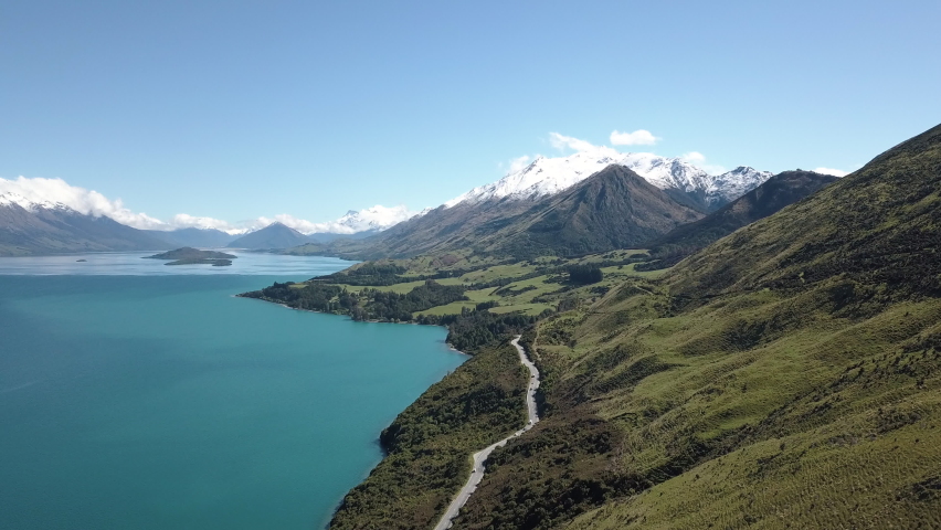Breathtaking aerial of crystal clear water on a paradise lake beside green rolling hills and a winding road through the mountains.