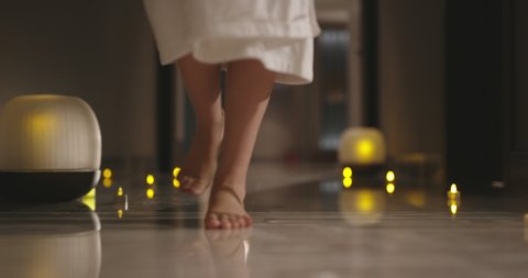 Close up shot of slender female legs slowly stepping on floor with candles. Woman in white robe walking in resort spa low angle 4k footage