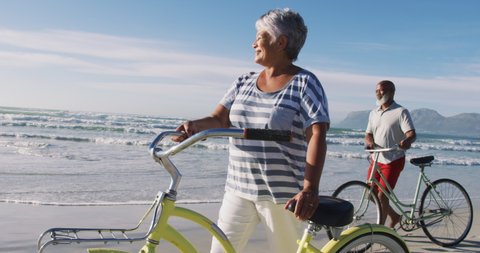 Smiling senior african american couple walking with bicycles at the beach. healthy outdoor leisure time by the sea.