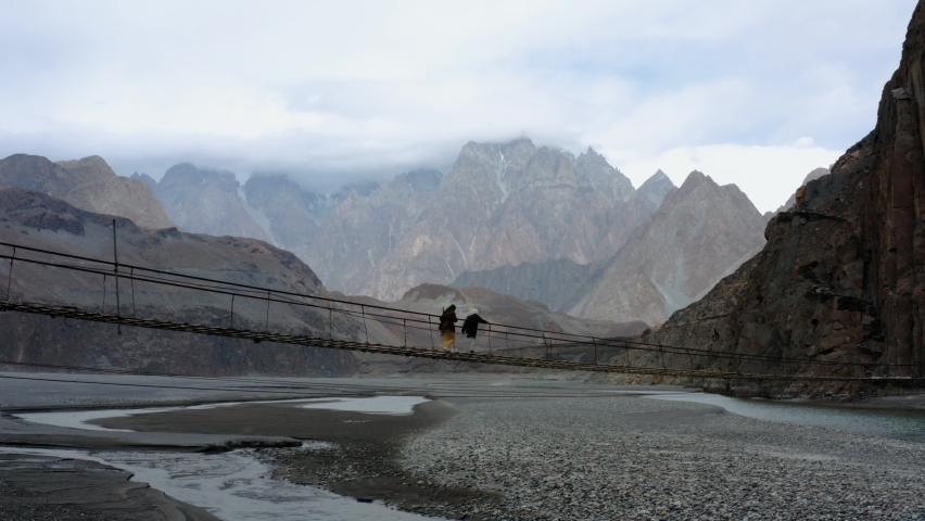 Women Walking Across The Suspension Bridge Of Hussaini With Rocky Mountain Ranges In Background At Hunza Nagar, Northern Pakistan. - Wide Shot