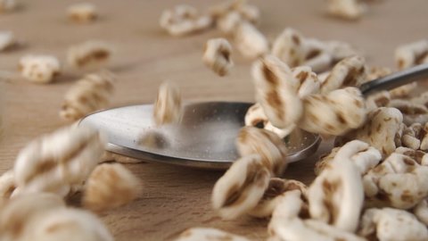 Uncooked cereal grains of puffed spelt wheat fall and fill a metal spoon close-up on a wooden cutting board in slow motion. Macro shot