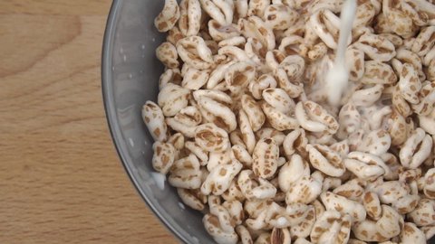 Pouring milk into a bowl with dry puffed spelt wheat in slow motion. Close up. Wooden background. Healthy breakfast cereal