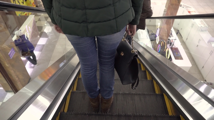 A girl goes down on an escalator in a shopping mall or supermarket. Women's feet in jeans, boots and a purse on the stairs go down in the store. Escalators and lifts in shopping centers, supermarkets Royalty-Free Stock Footage #1071488020