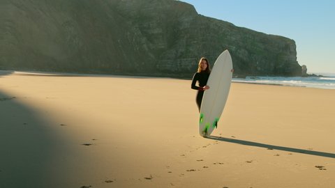 Beautiful cinematic shot of female surfer stand with professional surfboard deck on soft sand of epic beach. Soft light slow motion footage of woman surfer in wetsuit. Inspiring millennial lifestyle