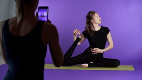 Flexible young woman is posing on the yoga mat, another woman is making video with the smarphone. Violet background
