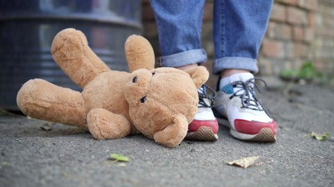 Close-up of teddy bear falling on asphalt at feet of young woman in sneakers. Unrecognizable Caucasian lady dropping toy outdoors in suburb slum