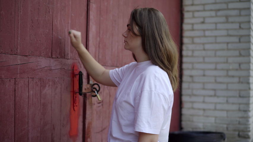 Side view of young beautiful Caucasian brunette woman knocking in big red door outdoors. Portrait of serious charming lady in industrial suburb on summer day
