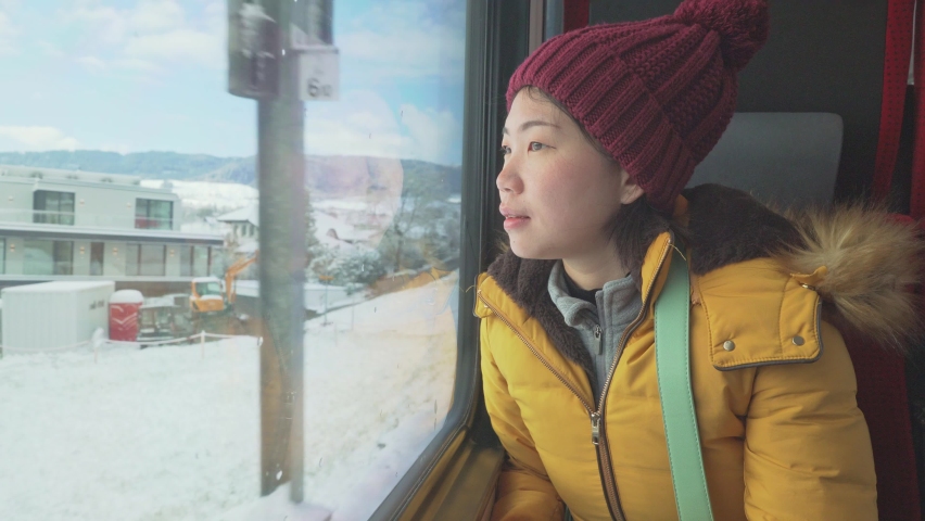 train travel getaway - young happy and beautiful Asian Japanese woman enjoying snow landscape through railcar window during Winter holiday trip
