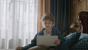 Child smiling and playing online by tablet lying on bed. Using handheld devices for entertainment. Communication via internet distantly. Boy having a video call at home.