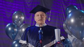 man in academic clothes with diploma in hands conducts ceremonial graduation ceremony and congratulates graduates into microphone background of balloons in assembly hall, look at camera