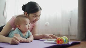 Happy Smiling Asian Young mother using laptop with her cute baby boy son lying on yoga mat at the floor together after practicing yoga and exercising. Loving Family at home. BeH3althy