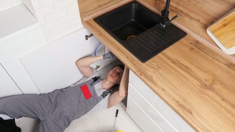 Skilled employee in grey uniform and gloves lies on floor and unscrews details repairing waste trap under sink in kitchen view from above